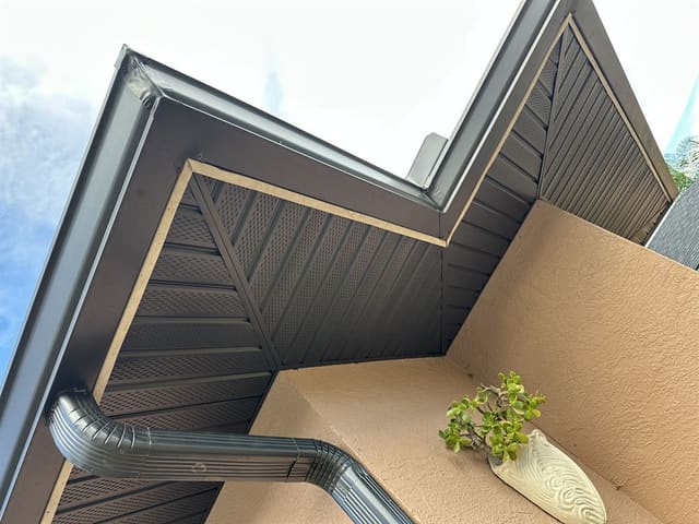 Bronze downspout with offset elbows running down a stucco wall under new vented soffit.