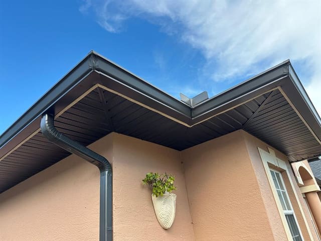 Bronze gutter, downspout and vented soffit meeting at a stucco house corner.